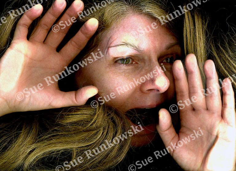 A close-up image of a woman’s face and hands pressed flat against a glass surface, creating a distorted effect. Her expression is one of alarm or distress, with wide eyes and an open mouth as if shouting or gasping. Her long, light brown hair fans out around her face, contributing to the sense of entrapment or urgency. The image conveys a raw, emotional intensity and a sense of being trapped or silenced.