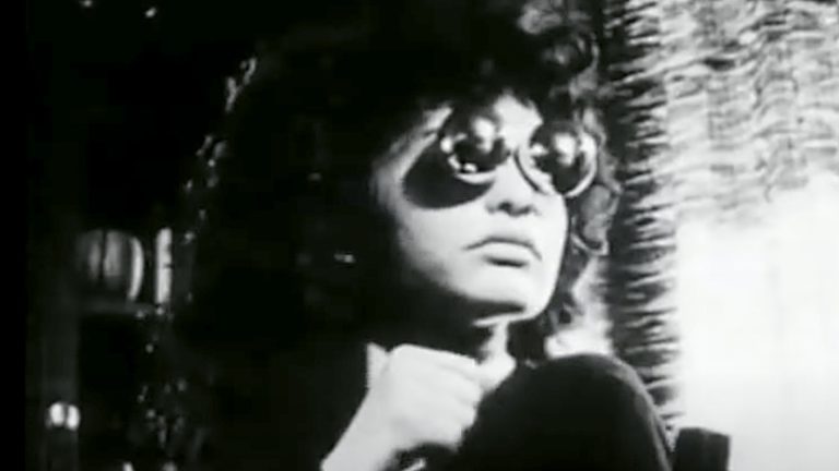 Black-and-white image of a person wearing large round sunglasses and a dark outfit, sitting indoors near a window with light streaming in. The person has curly hair and an introspective expression, with one hand raised near their chin, creating a moody, cinematic atmosphere.